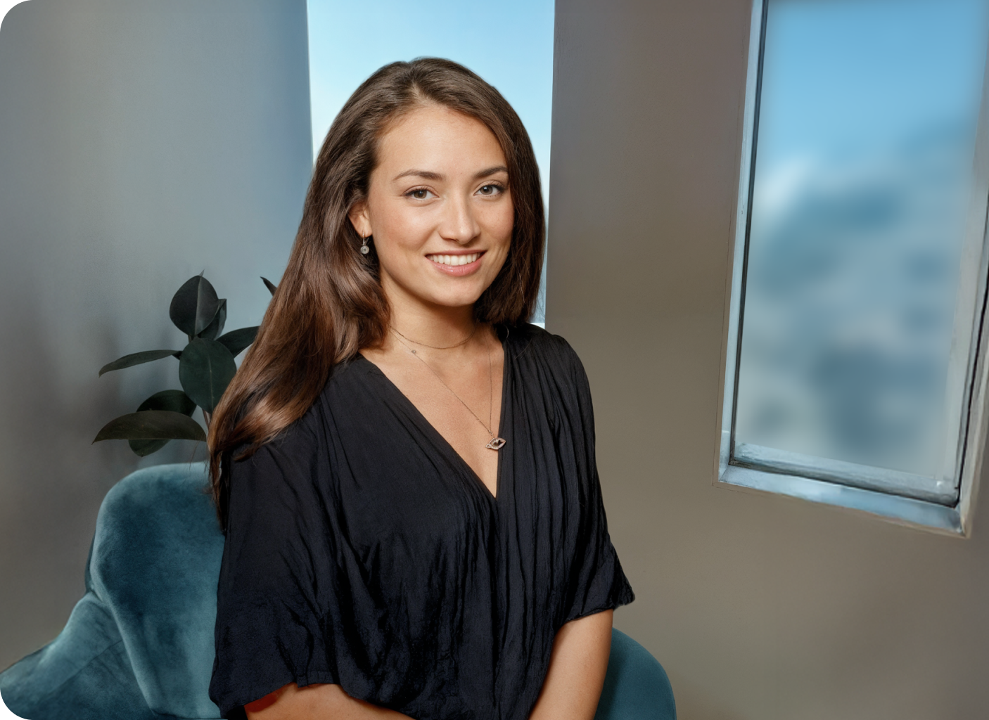 A woman with long brown hair, wearing a black blouse and necklace, sits on a blue chair near a window using a position template. A plant is in the background, and she is smiling at the camera in a well-lit room.