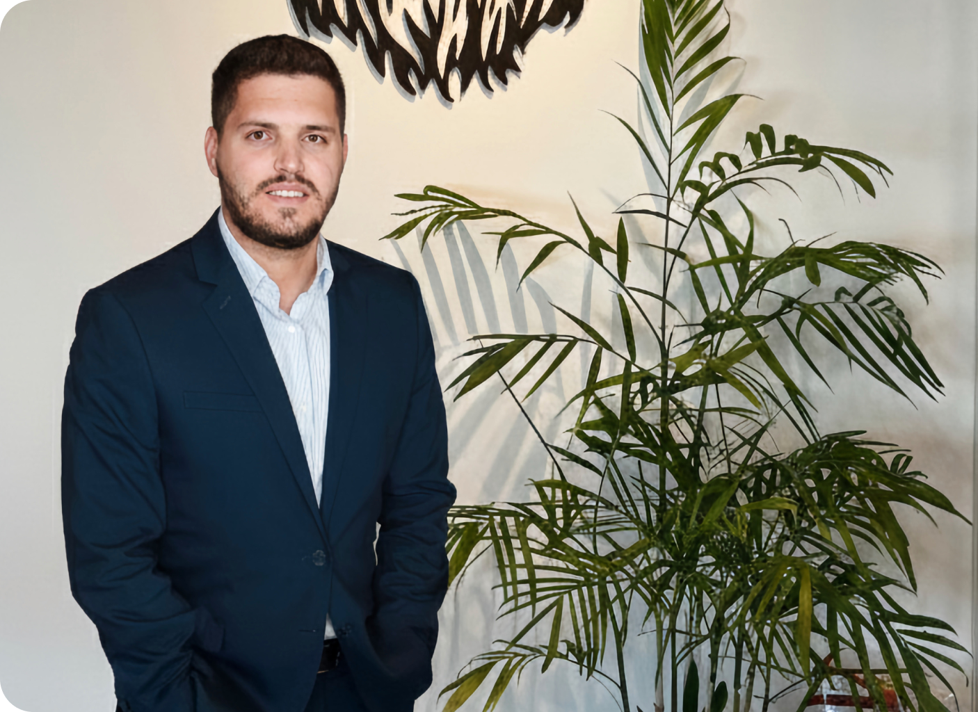 A man in a navy blue suit stands next to a tall green plant, with his hands in his pockets, in a well-lit indoor setting. A decorative wall piece and posiotion template1 are visible above him.