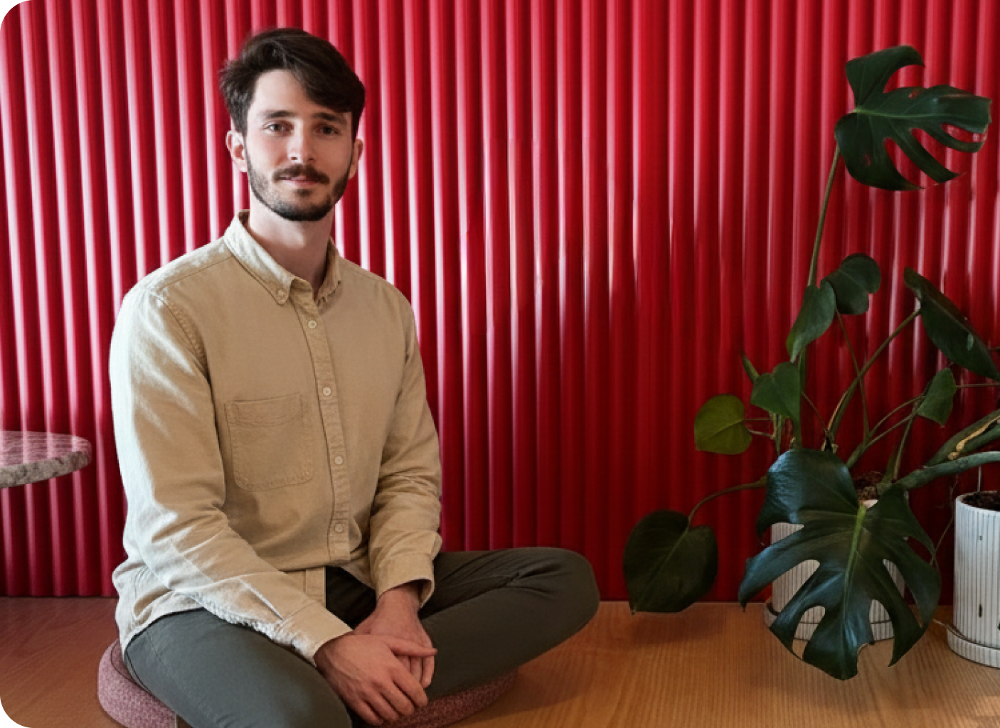 A man with dark hair and a beard sits cross-legged on a cushion in front of a red, ribbed wall, embodying the classic position template1. He wears a beige shirt and green pants, with a potted monstera plant to his right.