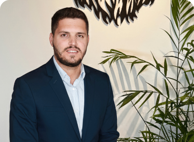A man with short dark hair and a beard, wearing a navy blue suit and light shirt, stands indoors next to a green leafy plant and a decorative wall piece, showcasing the posiotion template1 style.