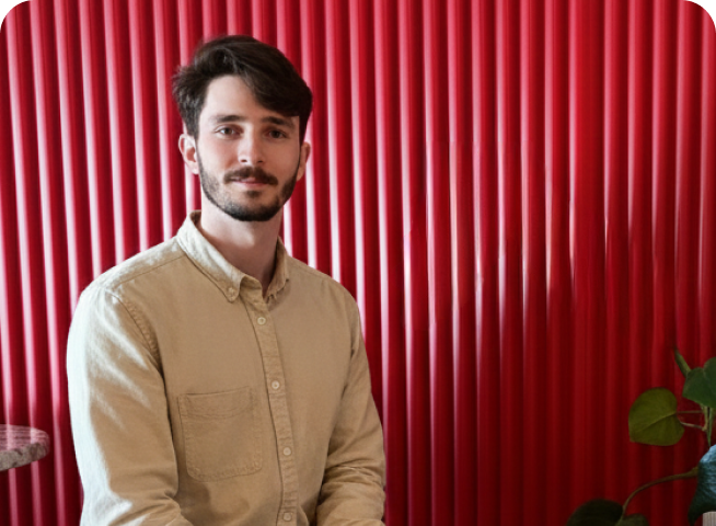 A young man with short dark hair and a beard, in a beige button-up shirt, sits in front of a red vertical panel wall following the position template1. A green leafy plant is partially visible on the right side.