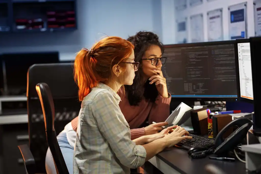Two women sit closely at a desk with multiple computer monitors displaying code. One types on the keyboard while the other looks at the screens, thoughtfully holding her chin. The room is softly lit and filled with tech equipment.
