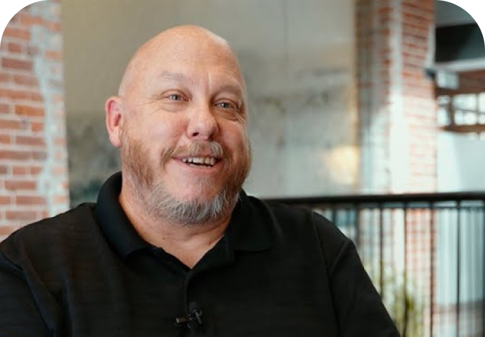 A smiling middle-aged man with a beard and a bald head, wearing a black collared shirt, sits indoors in a modern setting with exposed brick walls and large windows in the background.