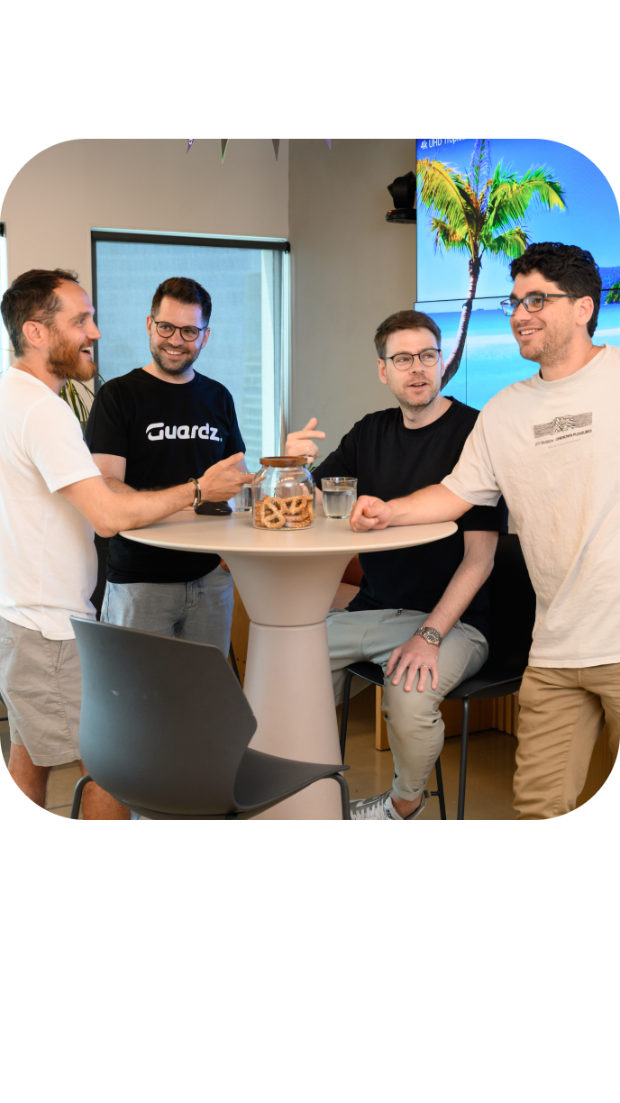 A group of people gather and chat around tables in a brightly lit, modern cafeteria decorated with triangular banners. There are plates of fruit and snacks on the tables.