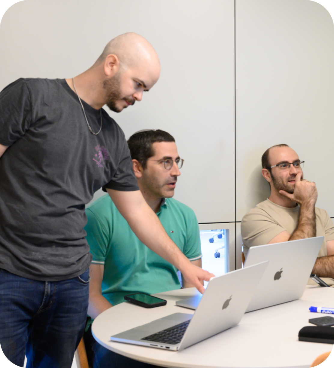 Two people sit across from each other at a small table with laptops, smiling and having a lively conversation in a modern workspace with wood-paneled walls and a dark circular sound panel.