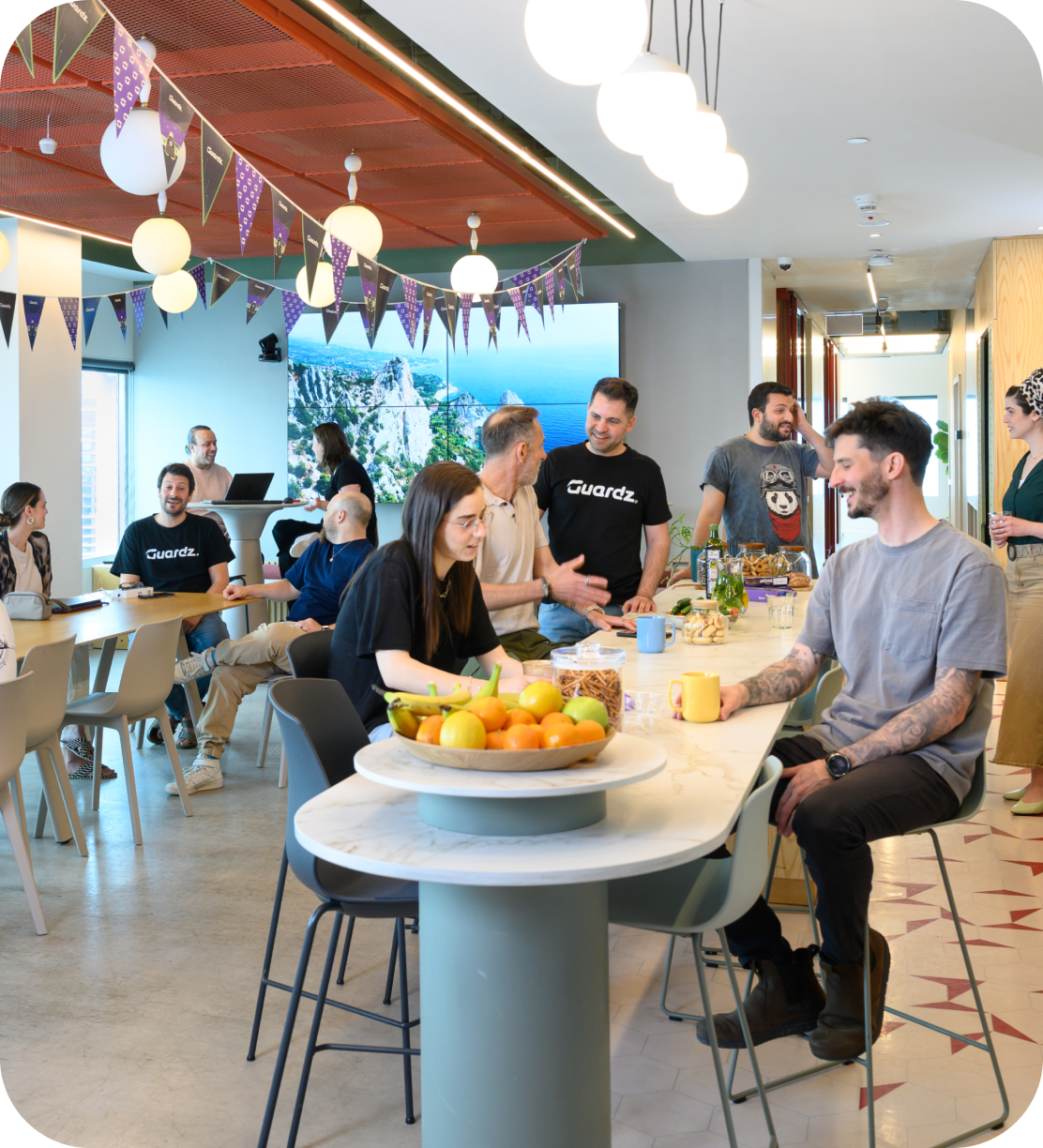 V2 Three men sit and stand around a table with a laptop, having a discussion in a modern office. One gestures while speaking, another listens thoughtfully, and the third looks on. A plant and a book are also on the table.