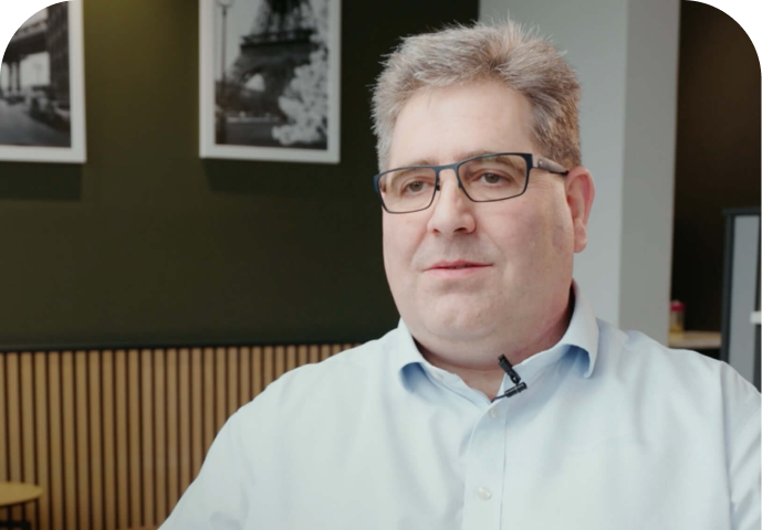 A middle-aged man with glasses and gray hair, wearing a light blue shirt, sits indoors with framed black and white photos and a wooden wall in the background. He has a microphone clipped to his collar.