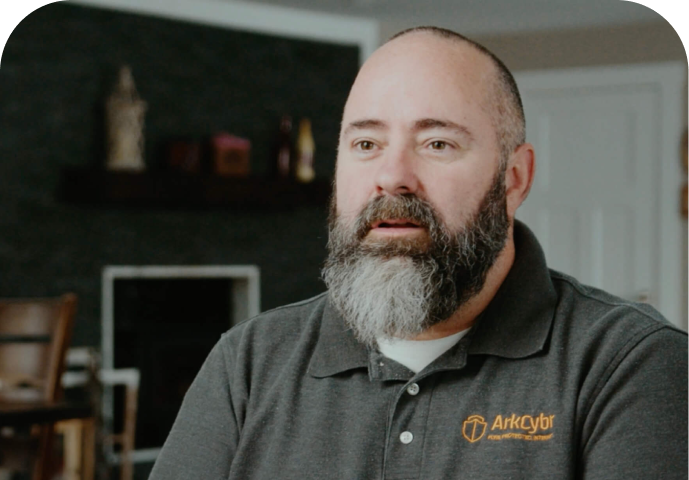 A bearded man wearing a gray ArkCyber polo shirt sits indoors, looking slightly to the side. The background includes a dark wall, a fireplace, and blurred furniture.
