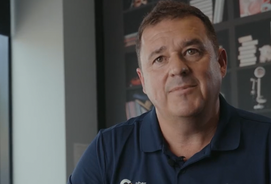 A man wearing a navy blue shirt sits indoors, looking slightly to the side. Shelves with books and decorative items are visible in the background.