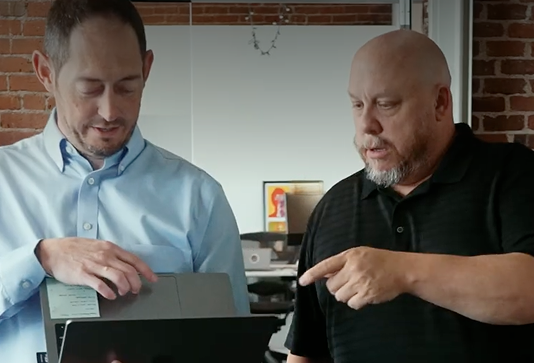 Two men standing indoors look at a laptop; one is holding and gesturing to the screen, while the other points at it, both appearing focused and engaged in discussion.