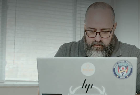 A bearded person with glasses works on a laptop covered in stickers, including an HP logo and a round badge, sitting in front of a window with blinds during the day.