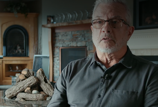 An older man with gray hair and glasses sits indoors, wearing a dark shirt with a microphone clipped on. Behind him are a stone fireplace, wooden logs, and a kitchen area with shelves and cabinets.