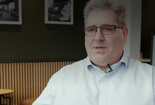 A man with short gray hair and glasses, wearing a light blue shirt with a lapel microphone, sits indoors against a wall with framed photos and wood paneling.