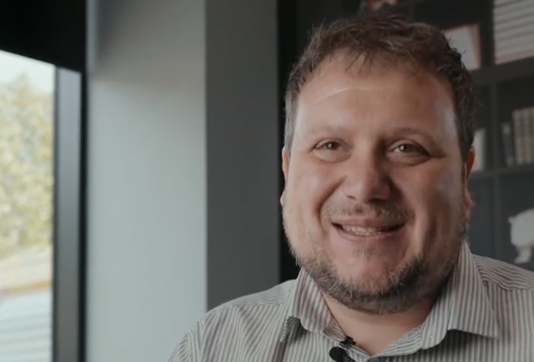 A man with short hair and a beard smiles at the camera in an office with a bookshelf and window in the background.