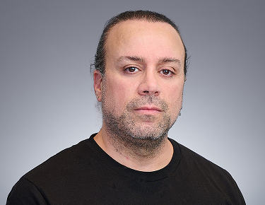 A man with long dark hair pulled back, wearing a black shirt, looks at the camera with a neutral expression against a plain gray background, resembling an S1 Simulation in style.
